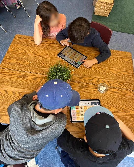 3 students sit at a table in pairs looking at shared iPads with grids visible on the screens.