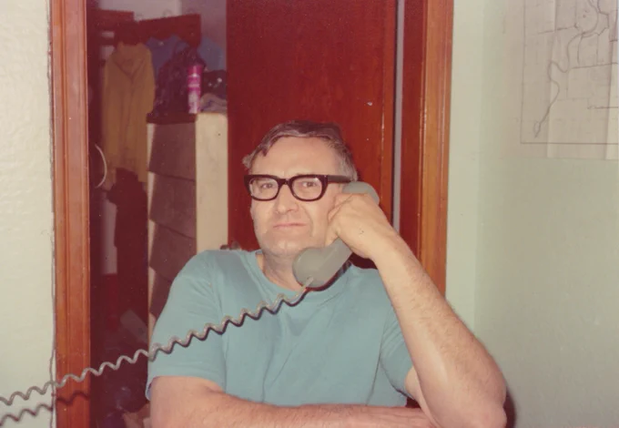The photo shows a person sitting in a room, holding a rotary phone to their ear. They are wearing a light blue short-sleeved shirt and glasses. The background includes an open closet with clothes hanging, a dresser with various items on top, and a wooden door. A map or drawing is pinned to the wall on the right. The coiled phone cord is prominently visible, suggesting the photo is from an earlier era, likely mid-20th century. The setting appears to be a casual, domestic environment.