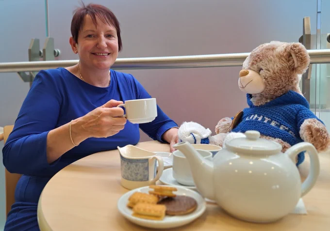 Woman holding tea cup sits at round table with teddy bear: teapot, cups and saucers, jug and saucer of biscuits in foreground