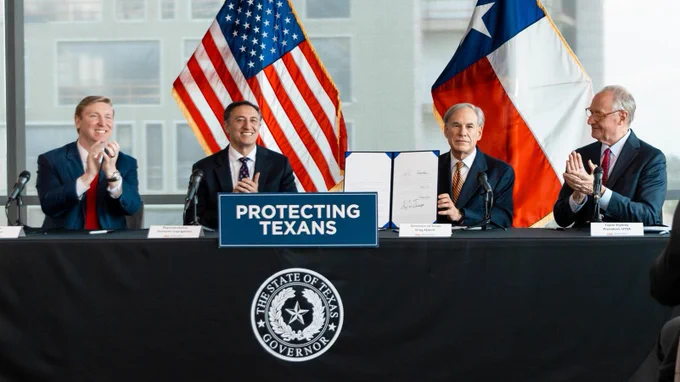 Four individuals (Left to right: Sen. Tan Parker, Rep. Giovanni Capriglione, Governor Greg Abbott, President Eighmy.) sitting at a table with a sign reading "Protecting Texans" and flags of the United States and Texas in the background. They appear to be involved in a formal event, possibly a signing ceremony.
