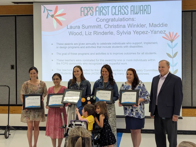Five women and one man are standing in front of a presentation screen with the title of FCPS First Class Award. The women are holding award plaques in front of them. Two students are also in front of the group and smiling.