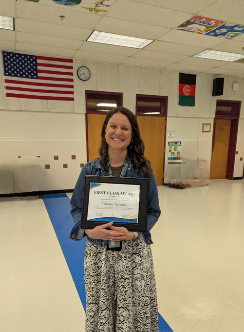 Christina is holding a First Class Award plaque and smiling in a cafeteria.