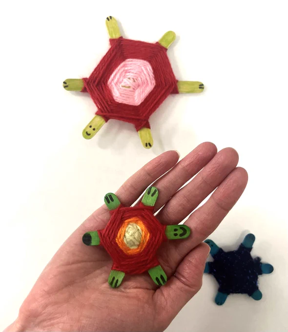 Photo of hand holding a children's craft turtle made from green lolly sticks and multi-coloured yarn, with two more handcrafted turtles on the pale surface below.