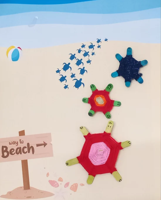 Photo of 3 children's craft turtles made from coloured lolly sticks and multi-coloured yarn, placed on a printed-out colour poster looking like a sandy beach with a sign reading "Way to beach" with a sea shore and and a multi-coloured beach ball in the background. The craft turtles have been placed to look like they are crawling towards the ocean.