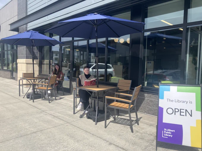 A yellow, green, blue, and purple St. Albert Library sign states The Library is Open. To the left are two wooden patio tables, both surrounded by three tables and covered with blue umbrellas. At each table sits a woman reading a book. 