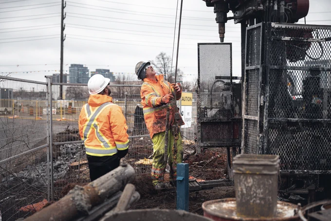 Two construction workers inspect equipment at a construction site, with buildings in the background.