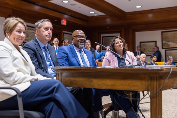 Governor Healey, Lieutenant Governor Driscoll, Secretary Tutwiler, and Secretary Gorzkowicz sit at a table in a formal hearing room, smiling and engaged in front of two microphones. In the background, an audience of people in suits listens attentively.