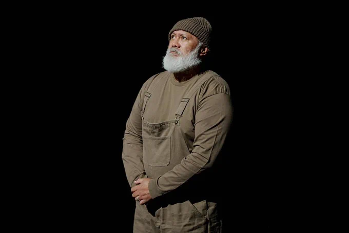 David Fane in a brown set of overalls and long sleeved shirt with a brown cap and white beard standing in the dark looking up at the light. 