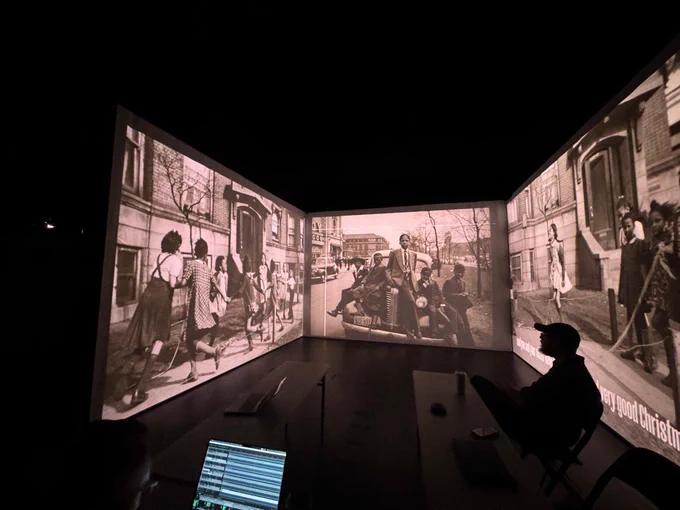 Three large screens featuring sepia-toned photographs from the early-to-mid 20th century. A man sits to the side looking at the screens. In the foreground is a laptop computer screen.