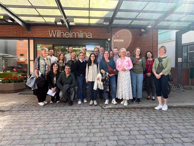 Fotografia de grup feta a l’exterior del centre Wilhelmiina, a Finlàndia. Hi apareixen una vintena de persones somrients, dretes davant de l’entrada de l’edifici, sota una marquesina de vidre. El fons mostra parets de maó vist i el rètol “Wilhelmiina” en lletres grans. El dia és assolellat i l’ambient acollidor.