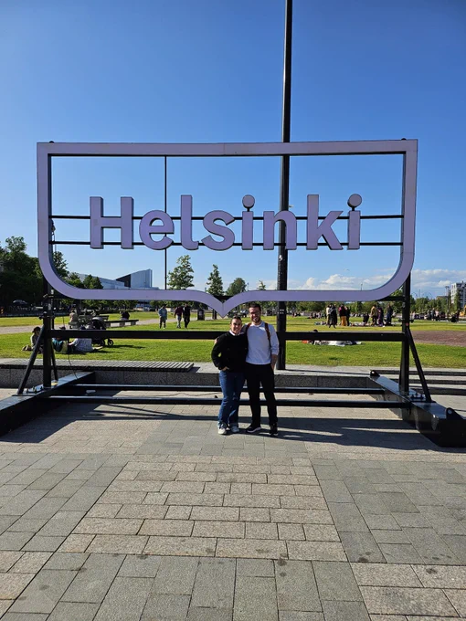 Dues persones somriuen davant d’un gran rètol metàl·lic amb la paraula “Helsinki” en lletres blanques. El cartell té forma de bombolla de diàleg i està situat en una plaça oberta i assolellada. Al fons, es veuen arbres, gespa i grups de persones assegudes o passejant, aprofitant el bon temps.  