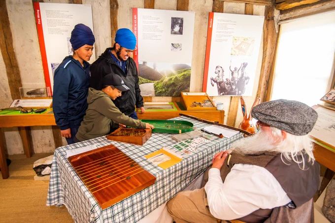 Two people wearing Sikh turbans stand behind a table with a child who is wearing a baseball cap. All three are smiling and looking at traditional games on a table. On the other side of the table sits a man wearing a flat cap. In the background are display panels with text and photographs on them, mounted on wattle and daub walls.
