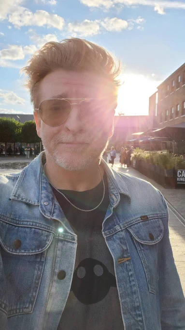 Rhys darby standing in the UK with the sun behind him doing a lense flare. Wearing a Jean Jacket and a black shirt, and a silver chain. His hair is looking floofy and unfairly hot as normal