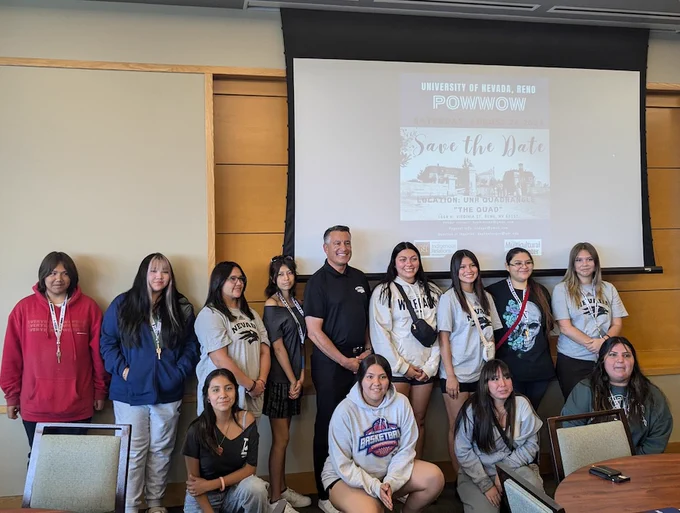 Group of people posing in a conference room with a poster for the University of Nevada, Reno Powwow event in the background.