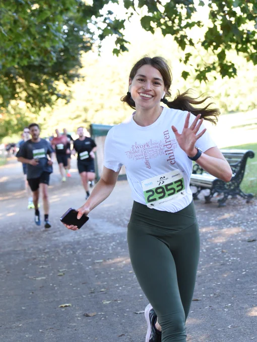A smiling lady waving to the camera, running in a white African Children's t-shirt and leggings. She has race number 2953 pinned to her t-shirt and is running under a tree with other runners in the background.
