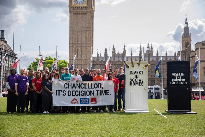 Campaigners from Shelter, National Housing Federation, Pathway, Action for Children, Single Homeless Project, St Mungo's, Centrepoint, Homeless Link, Big Issue, Amnesty International UK and Health Equals stand in front of two Parliament, next to two doors. 

One door, white and surrounded by arrows pointing towards it, has 'INVEST IN SOCIAL HOMES' written in large black font. 

The other door, next to it, is black and has 'WORSEN THE HOUSING EMERGENCY' in white font. 

The campaigners hold a sign which says in large letters 'CHANCELLOR, ITS DECISION TIME'.