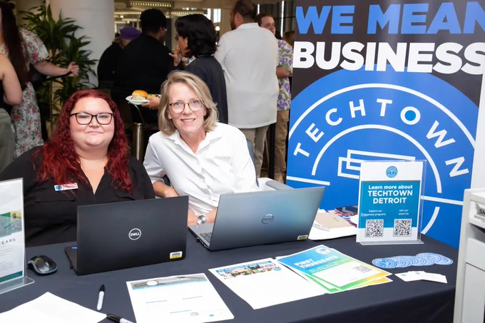 Two individuals seated at a TechTown Detroit promotional table with laptops, brochures, and a banner reading "WE MEAN BUSINESS" in the background.