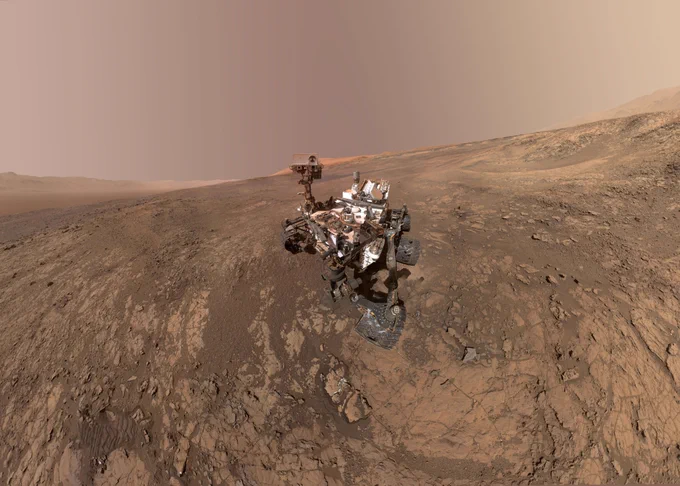The Curiosity rover sits on a rocky, sloped terrain on Mars, captured in a wide-angle selfie. The rover’s robotic arm is visible, and its body is covered in dust from years of exploration. The landscape stretches out behind it, featuring layered, reddish-brown rock formations, scattered boulders, and a soft, dusty sky. In the distance, gentle ridge lines and sedimentary slopes of Mount Sharp are visible under a pale Martian haze.