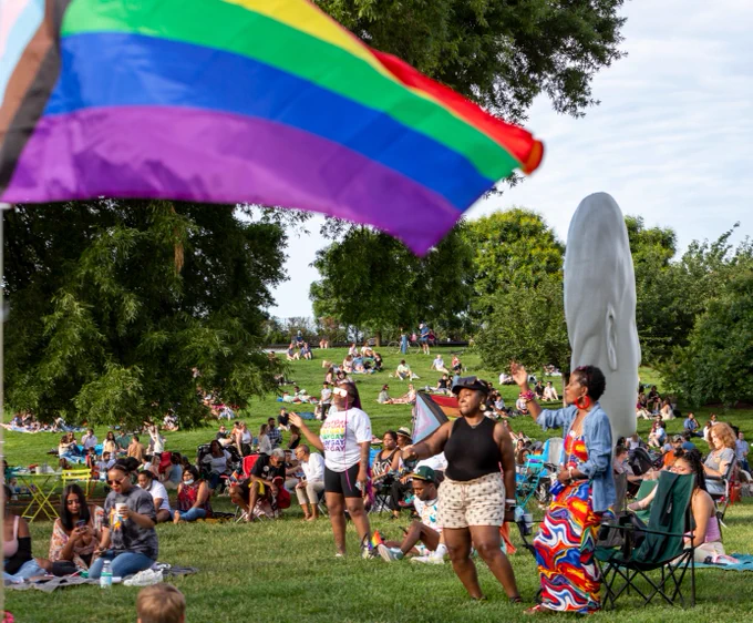A large group of people gather outdoors in VMFA's Robins Sculpture Garden. A large Pride Progress flag waves in the foreground.