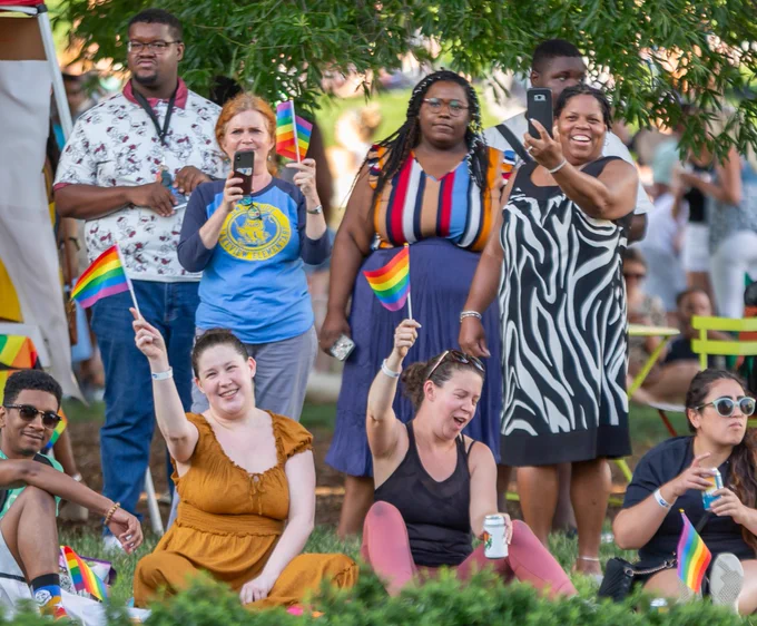 A group of people smile and pose outdoors. Some wave small Pride flags.