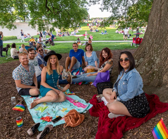 A group of friends sit posing at an outdoor event in VMFA's Robins Sculpture Garden. They are surrounded by a few pieces of Pride-themed rainbow memorabilia.