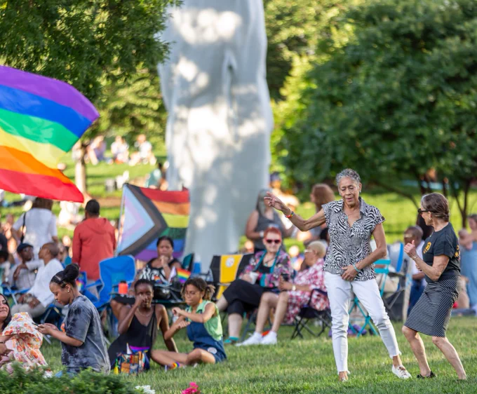 Two individuals dance at a crowded outdoor event in VMFA's Robins Sculpture Garden. Pride flags wave in the foreground and background.