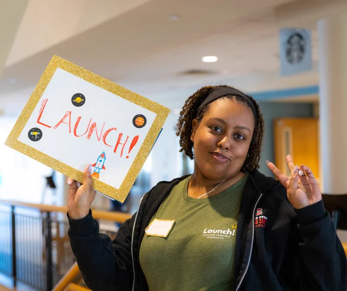 Person holding a sign that reads "LAUNCH!" with drawings of planets and a rocket, making an OK gesture, inside a room with a Starbucks logo visible in the background.