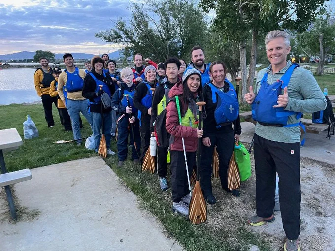 a group of people with life jackets holding oars in front of a lake next to some trees on one side and a table on the other.