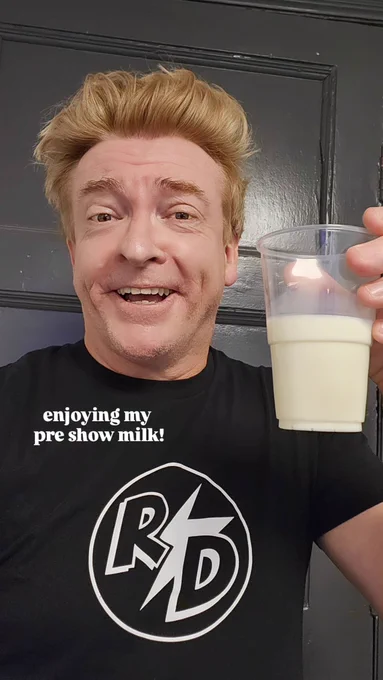 Rhys Darby in his RD shirt holding a glass of milk with white text that says "enjoying my pre show milk!"