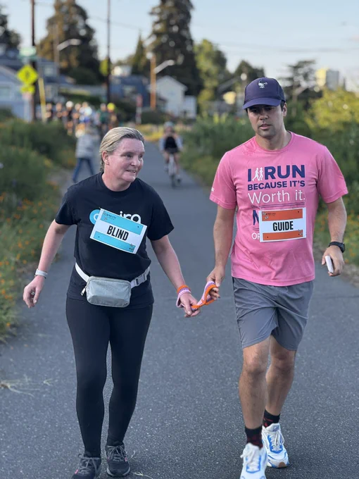 Two people running together outdoors on a paved path. The woman on the left is wearing a black t-shirt with a large sign on her chest that says "BLIND" in bold letters, black leggings, and black running shoes. She has a light gray waist pouch and is holding a short orange tether in her right hand. The man on the right is wearing a pink t-shirt that reads "I RUN BECAUSE IT'S Worth it." and a sign on his chest that says "GUIDE." He is also wearing gray shorts, white and blue running shoes, a dark cap, and a watch. They are holding the orange tether together, indicating he is guiding her. Both appear to be smiling and in motion, with greenery and flowers visible along the side of the path.