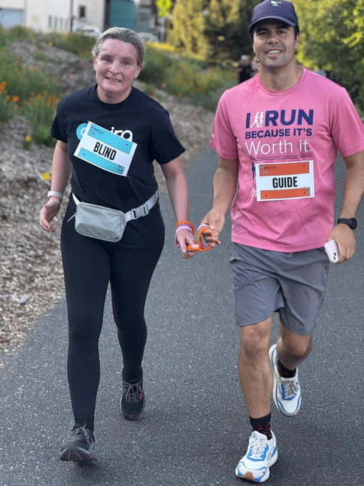 A woman with a "BLIND" bib and a man with a "GUIDE" bib are running together, holding a short orange tether, on an outdoor path.