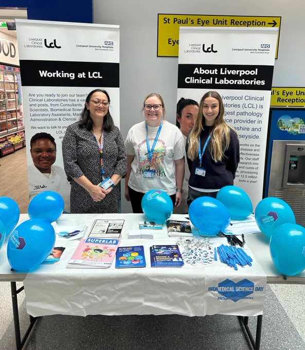 Three LCL colleagues smiling at the camera. the three are stood behind a table that is covered in various marketing materials: leaflets, booklets, pens, and balloons. Behind them is two LCL information stands. 