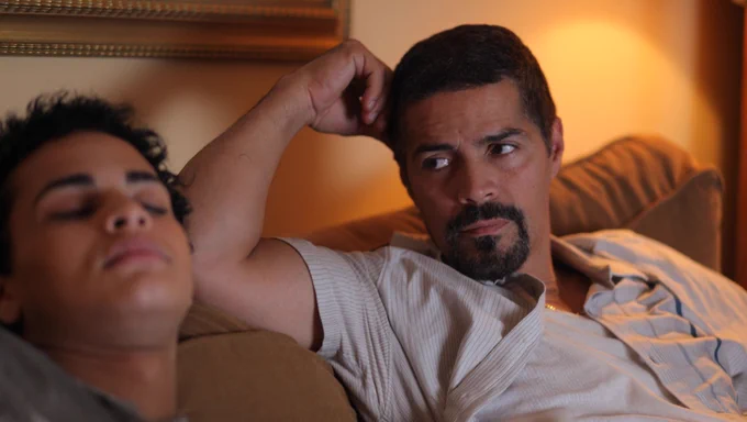 The actor Esai Morales sits on a couch, leaning his head on his hand, looking at his trans daughter who is lying down with eyes closed.