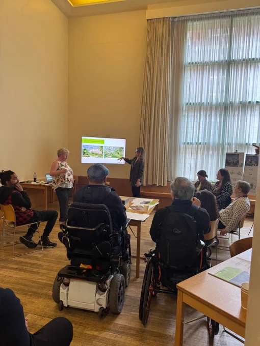 A photo of a presentation being delivered to a mixed group of adults in a communal meeting room.