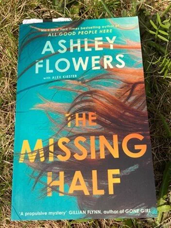A paperback copy of the novel The Missing Half by Ashley Flowers, co-written with Alex Kiester, lies on green grass in bright sunlight. The cover features long red hair swirling underwater against a teal-blue background, with the title in bold yellow and orange letters. A blurb at the bottom praises it as “A propulsive mystery” by Gillian Flynn, author of Gone Girl.