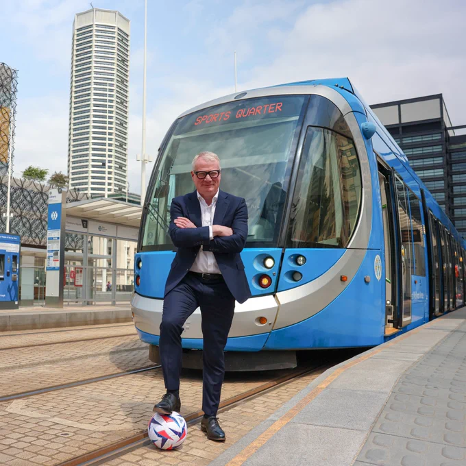 Photograph of a man with a football under his foot, stood behind a tram. 