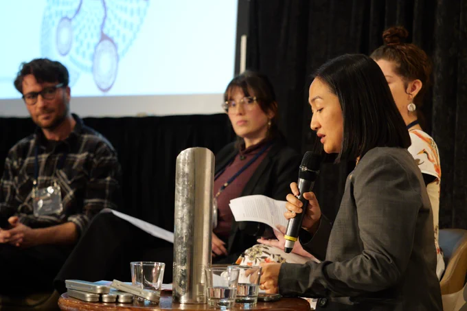 Three people look to a fourth who is speaking in to a microphone on a stage.