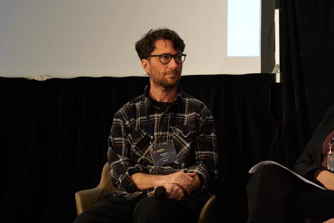 A man with facial hair and glasses wearing a black flannelette shirt looks to his left.