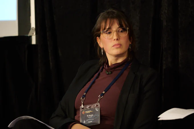 A woman wearing glasses and a maroon top leans back in her chair.