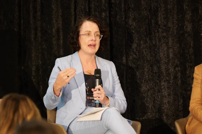 A woman in a brown blazer and orange glasses is smiling while holding a microphone.