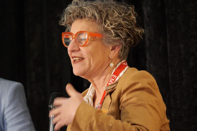 A woman with curly brown hair in a blue suit speaks into a microphone.