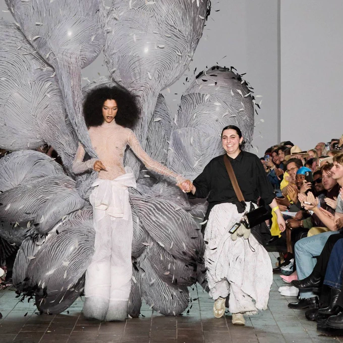 Mayah Hasbany walks the runway holding hands with a model wearing a dramatic, sculptural feathered outfit.