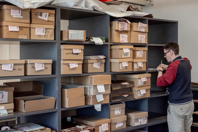 A staff members writes down notes while stood next to a large shelving unit stacked with dozens of labelled cardboard boxes.