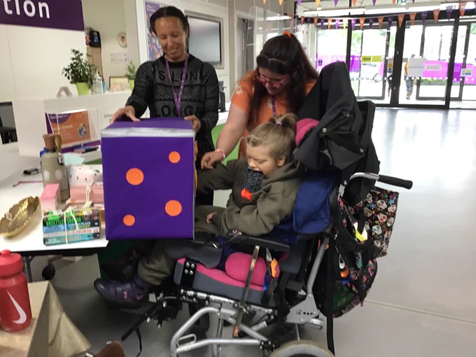 A group of three women including one sitting in a wheelchair picking a raffle ticket number out of a purple and orange spotty box. 