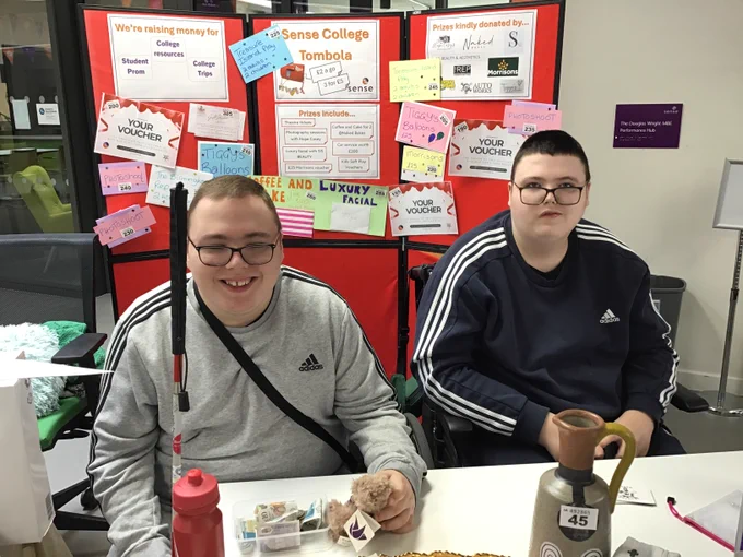 Two young men sitting at a table. They're both wearing glasses and Adidas sweatshirts. Behind them is a red display board covered in posters advertising the 'Sense College Tombola'. 