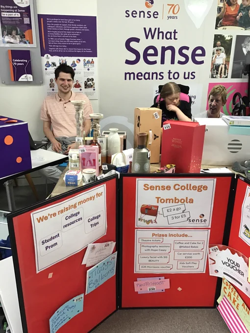 A young man wearing a pale red polo shirt and woman wearing a black top sit behind a table filled with tombola prizes. There are candle stick holders, bottles of perfume, vases, bottes of wine and more, all with a number stuck to them. In front of the table is a display board with information about the 'Sense College Tombola'. 