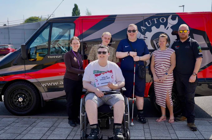 A group of people including one person sitting in a wheelchair standing in front of a van, branded with grey and red graphics. 