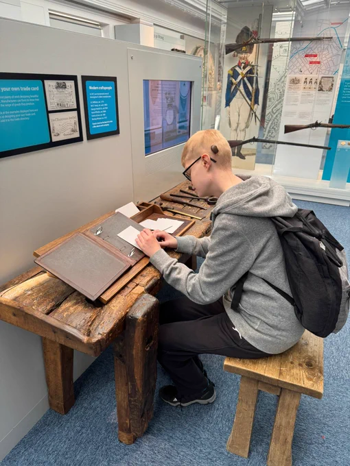 A young man wearing a cochlear implant sitting as a desk, drawing on a piece of paper. 