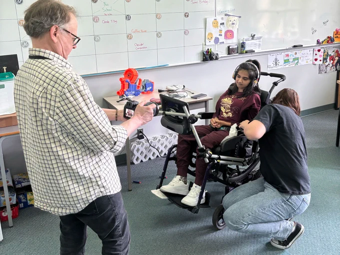 A man on left hold camera pointing towards a student in a wheel chair. Beside her a woman crouches to adjust something that appears to be on her chair.