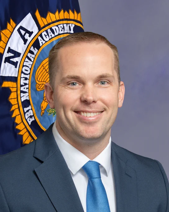 Official portrait of an individual smiling, wearing a business suit, in front of the FBI National Academy flag.